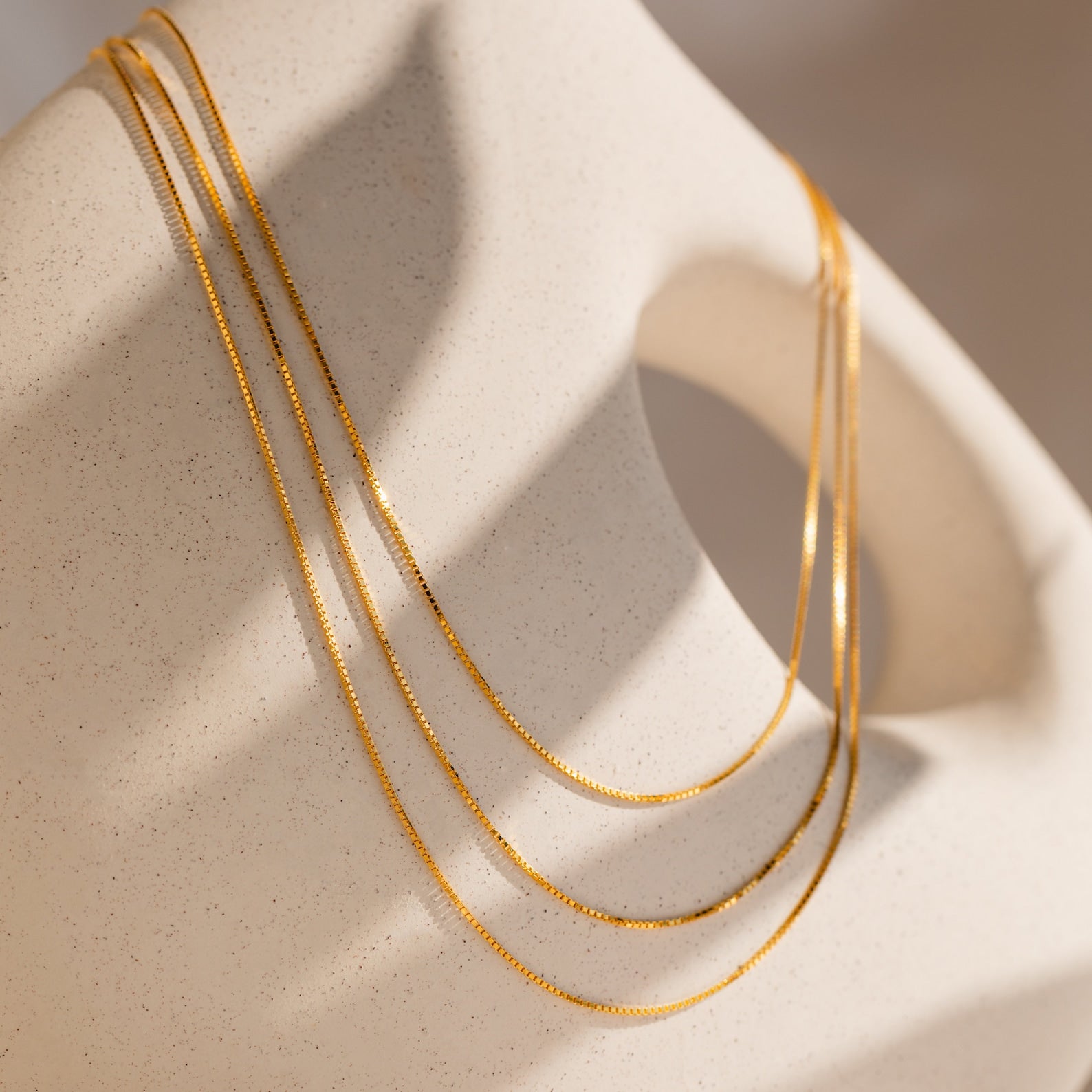 The Triple Box Chain Necklace, featuring three delicate gold layers, drapes elegantly over a smooth, abstract beige sculpture under soft lighting.