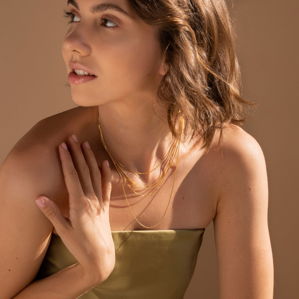 Woman in a strapless olive dress wears the Triple Box Chain Necklace set, showcasing layered necklaces as she looks to the side with one hand on her chest.