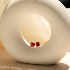 Emerald Birthstone Studs: gold gemstone earrings featuring square red stones, displayed in a round white ceramic sculpture on a beige surface—an elegant, personalized gift idea.