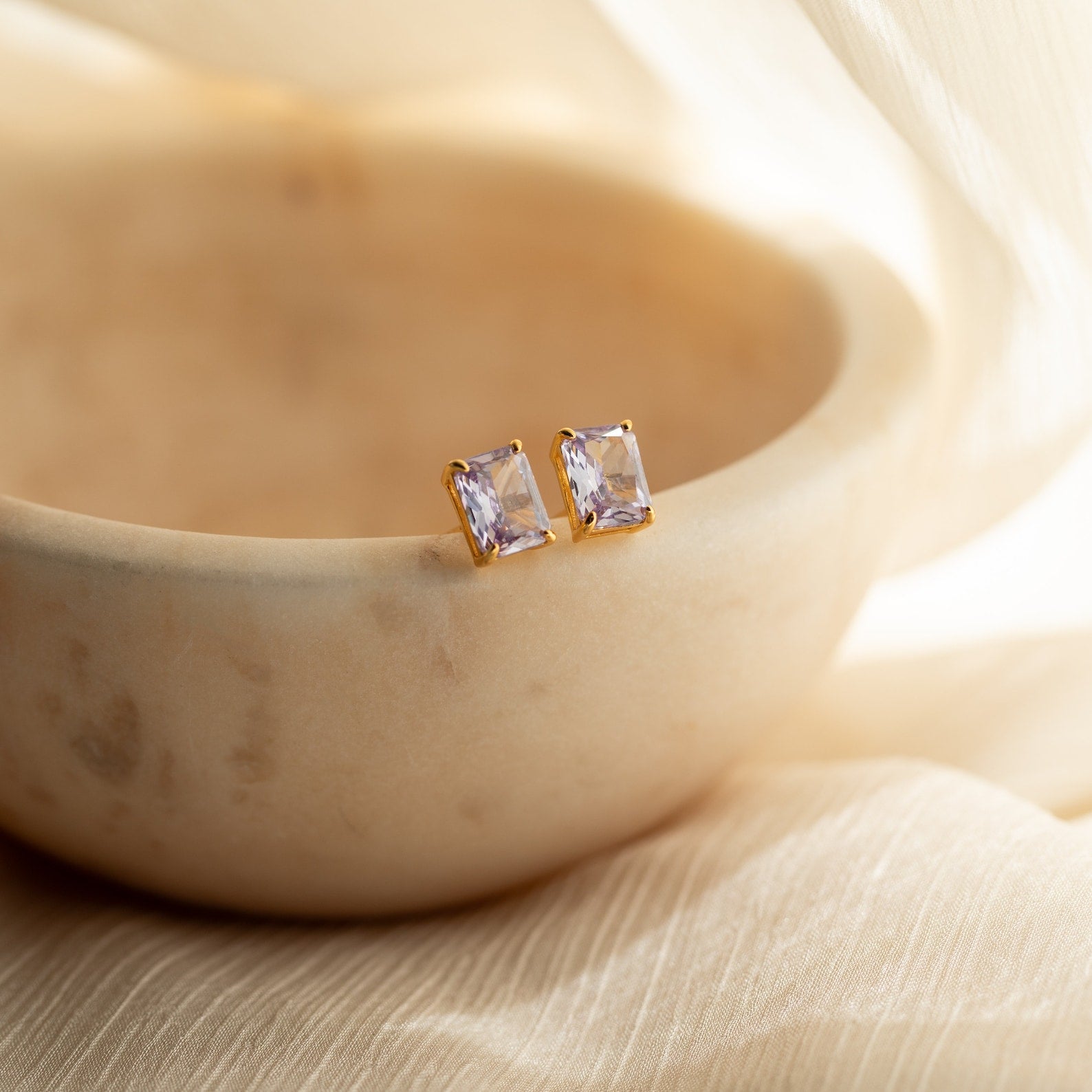Emerald Birthstone Studs featuring gold settings and rectangular clear stones rest elegantly on the rim of a smooth beige bowl.