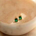 Emerald Birthstone Studs with rectangular green gemstones rest in a beige ceramic dish, bathed in sunlight. These elegant gemstone earrings make perfect personalized gifts for any occasion.