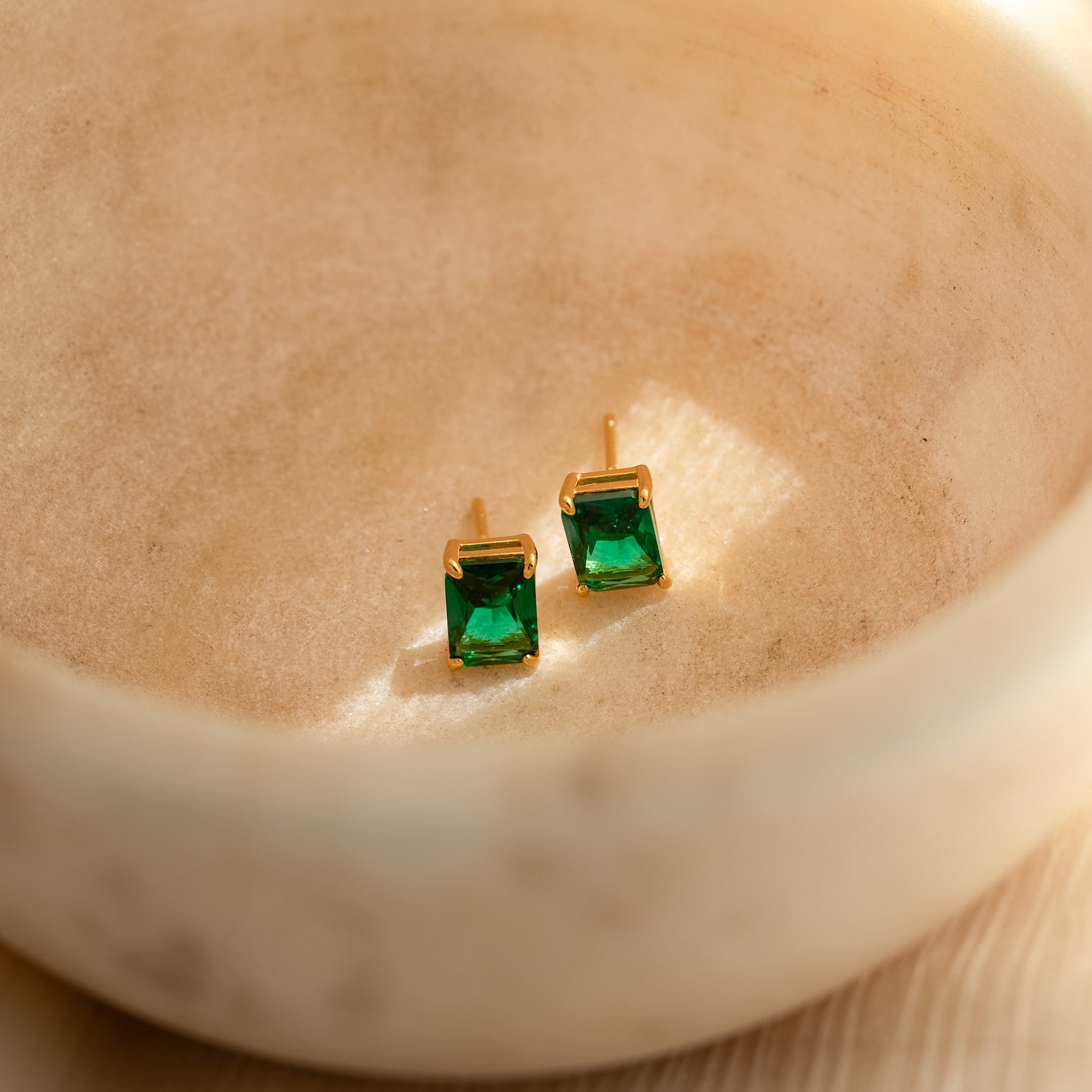 Emerald Birthstone Studs with rectangular green gemstones rest in a beige ceramic dish, bathed in sunlight. These elegant gemstone earrings make perfect personalized gifts for any occasion.