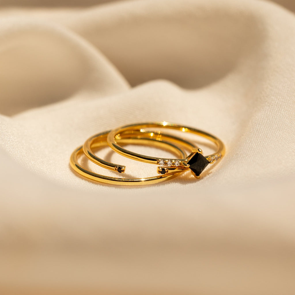 The Black Abyss Ring Set features three gold dainty bands adorned with black gemstones and small diamonds, shown on a soft beige fabric background.
