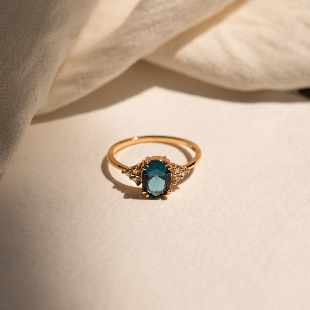 The Elizabeth Blue Topaz Ring features a vibrant blue gemstone and small clear stones, beautifully showcased against a soft, light-colored fabric background.