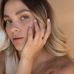 A woman with wavy blond hair touches her face, wearing the Elizabeth Blue Topaz Ring in 18K Gold, featuring a striking dark blue gemstone, set against a neutral background.