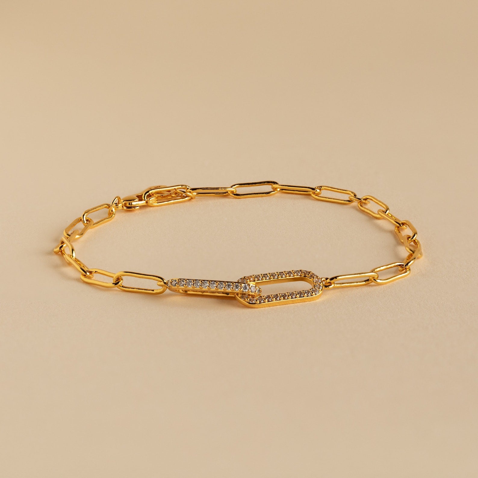 The Pave Link Chain Bracelet features elongated gold links and a crystal-encrusted clasp, making it an eye-catching statement piece, shown here on a beige background.