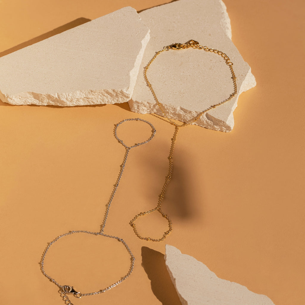 The Beaded Hand Chain Bracelet pairs effortlessly with delicate silver and gold chain bracelets, all displayed on beige stones against a tan background for a chic statement look.