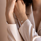 A person wears a cream blouse and the Carrie Initial Bracelet with a heart charm, elegantly showcasing personalized jewelry on their wrist.