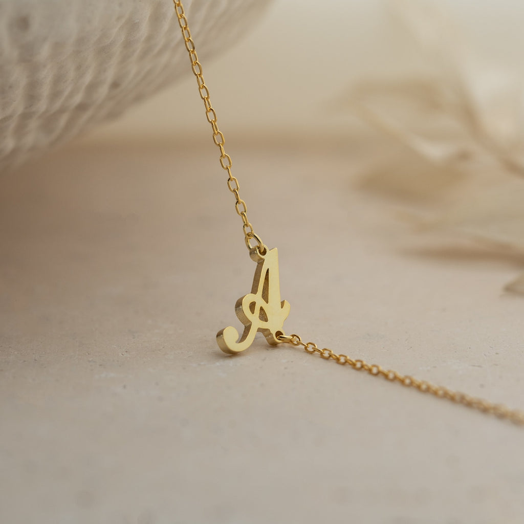 The Carrie Initial Bracelet features a cursive letter A pendant, displayed on a neutral surface—perfect for pairing with a delicate gold necklace or other personalized jewelry.