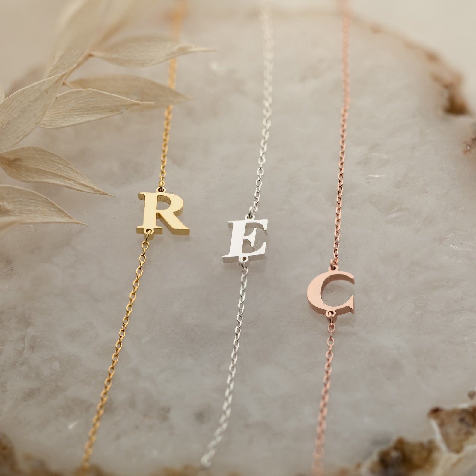 Three Newsletter Initial Bracelets in gold, silver, and rose gold, each with a personalized letter charm—R, E, or C—adorn a marble surface for a custom touch to your style.