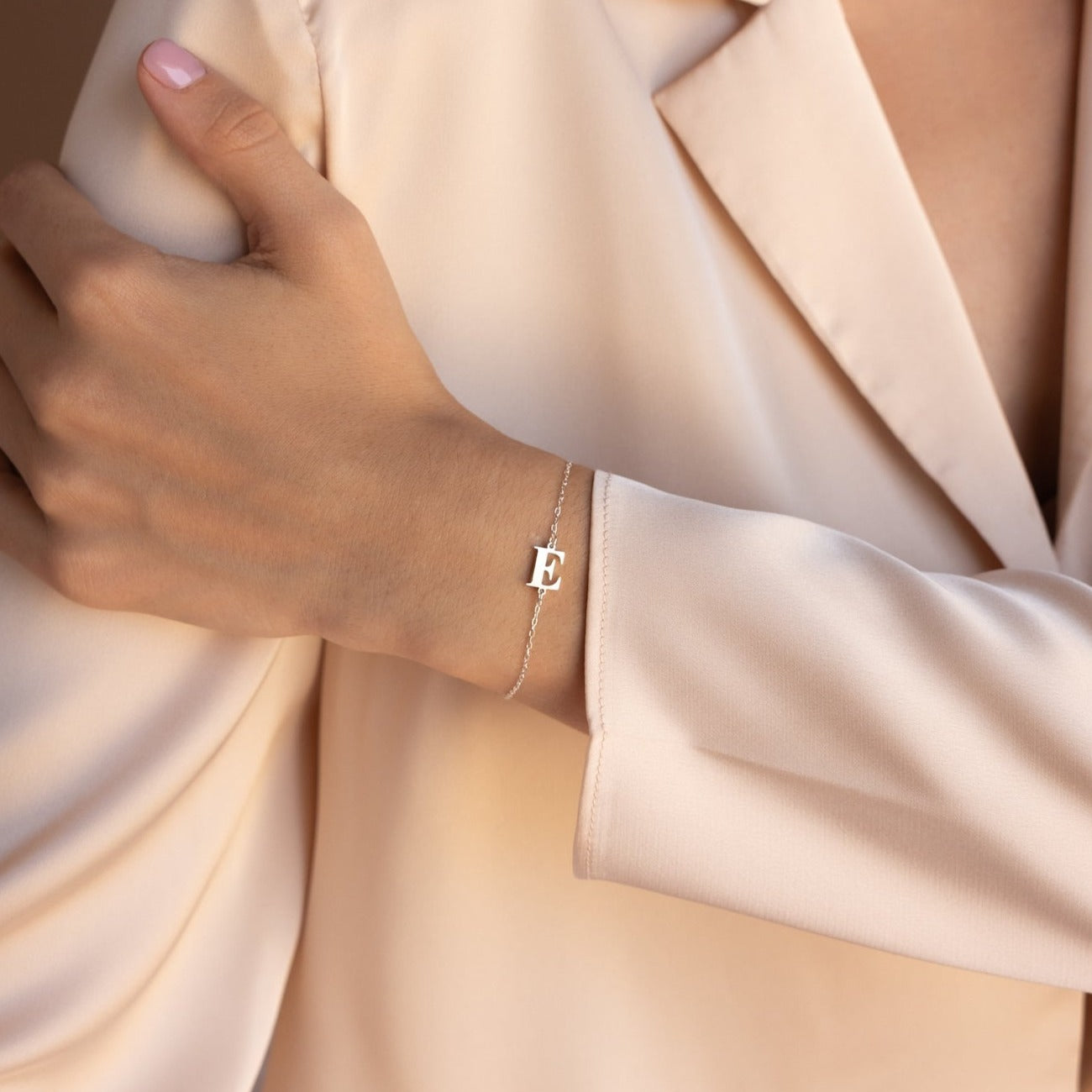 A person wearing a beige blazer and the Newsletter Initial Bracelet—a delicate, personalized bracelet with an E charm.