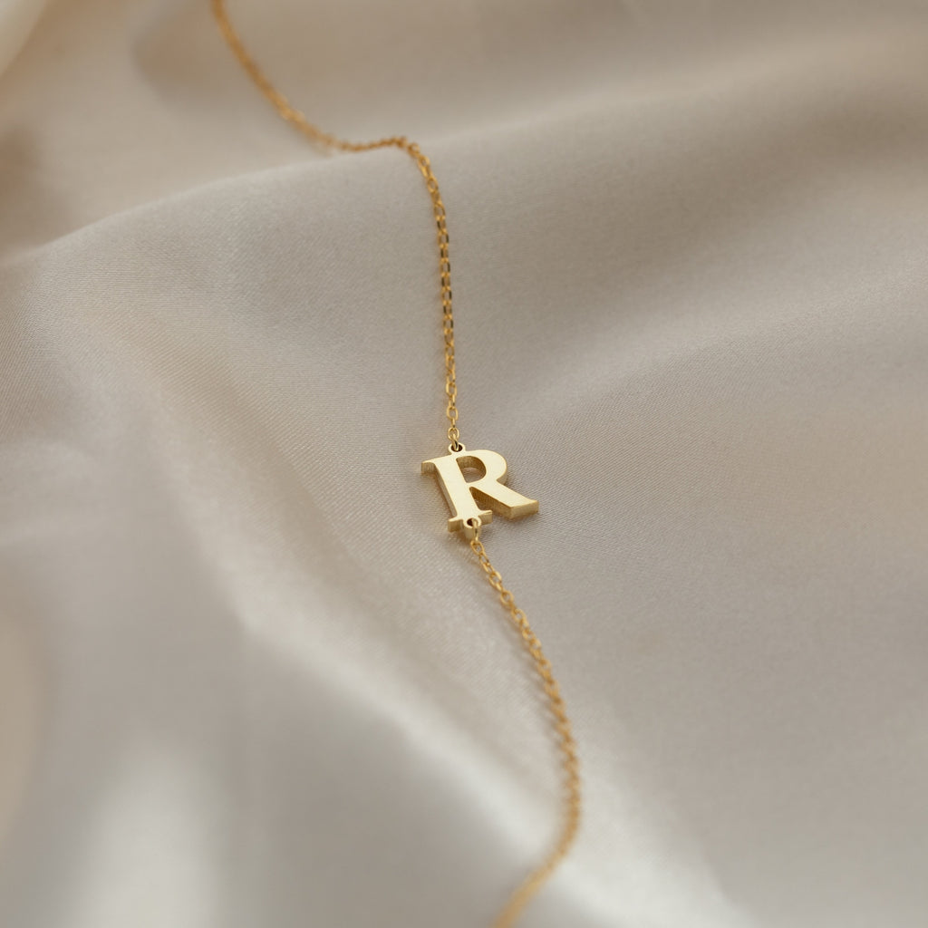 The Newsletter Initial Bracelet features a letter R charm on smooth, ivory-colored fabric—ideal for pairing with a gold necklace or another delicate bracelet to create a unique, personalized style.