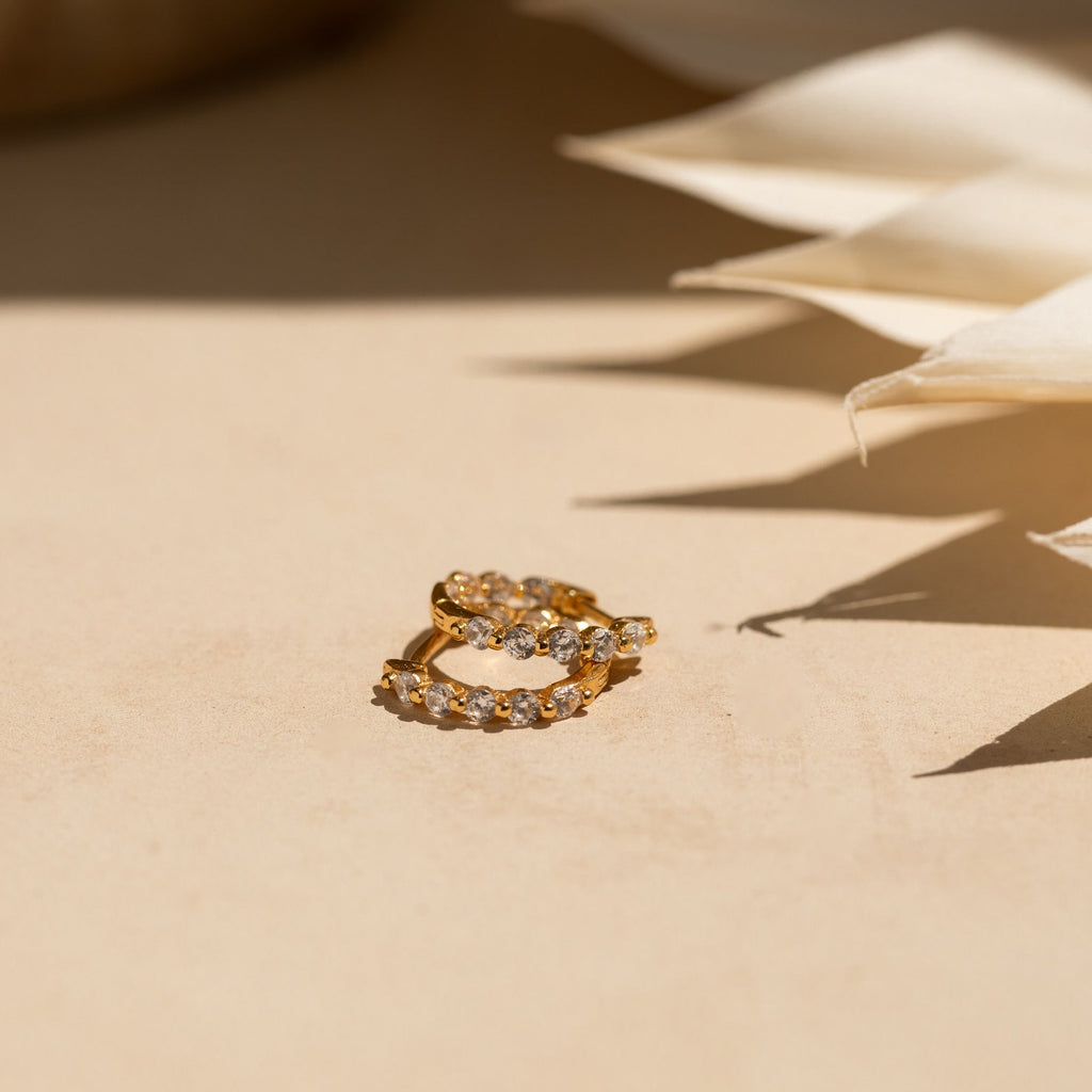 Inside Out Diamond Huggies in 18K Gold are displayed on a beige surface, with soft shadows and fabric nearby enhancing the elegant sparkle of their shimmering diamonds.