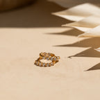 Inside Out Diamond Huggies in 18K Gold are displayed on a beige surface, with soft shadows and fabric nearby enhancing the elegant sparkle of their shimmering diamonds.