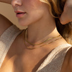 Layered gold necklaces with sleek chain designs, worn by a woman in a beige knit top.
