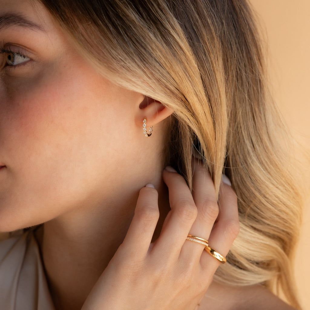 A woman touches her blonde hair, wearing Inside Out Diamond Huggies in 18K Gold and two gold rings, set against a beige background.