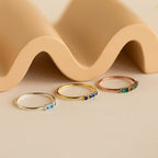 The Morse Code Birthstone Ring, a delicate colored gemstone ring, is displayed against a wavy beige background.