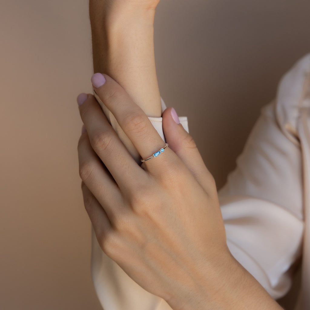 A hand with light pink nails wears a delicate Morse Code Birthstone Ring featuring blue stones, resting on a beige sleeve.