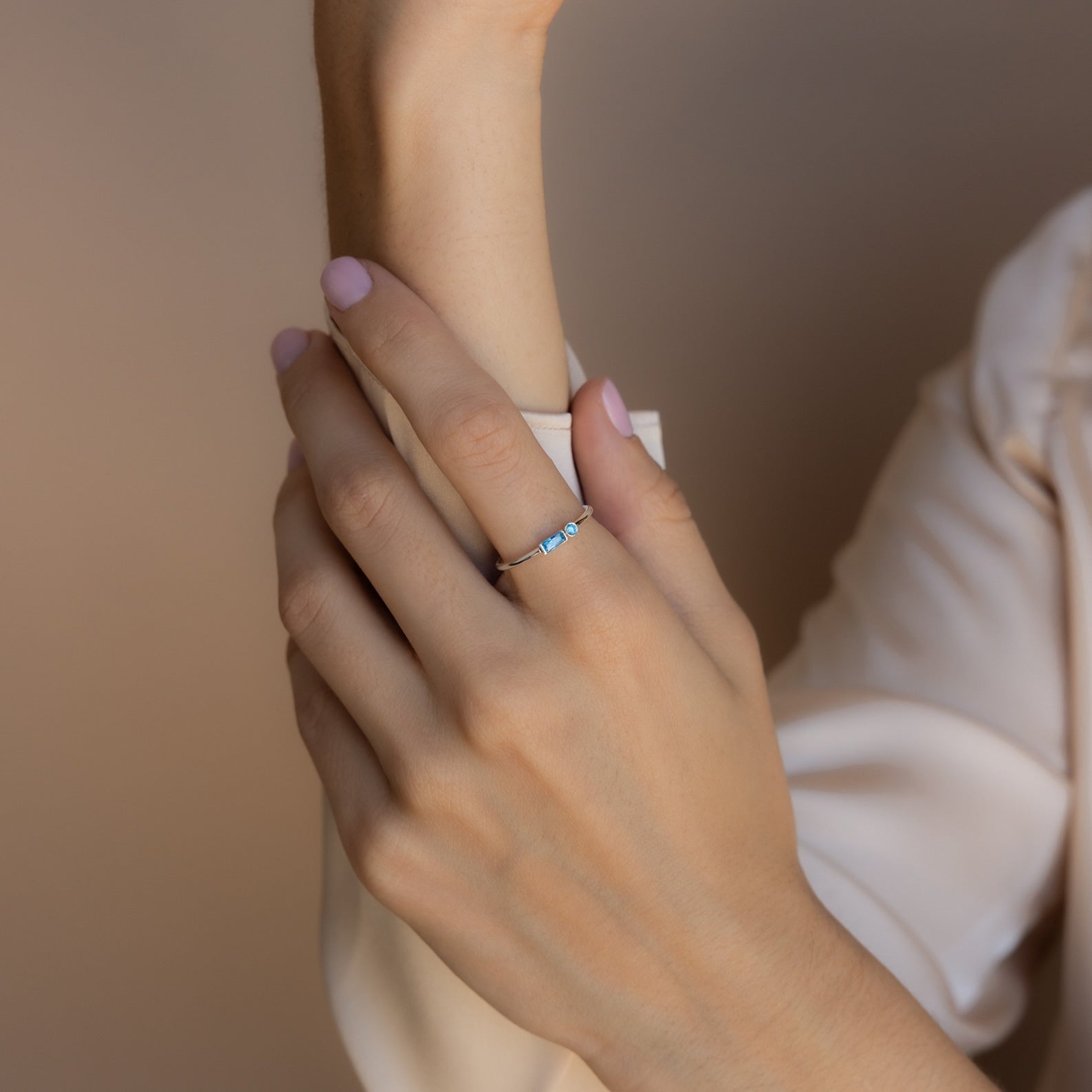 A hand with light pink nails wears a delicate Morse Code Birthstone Ring featuring blue stones, resting on a beige sleeve.