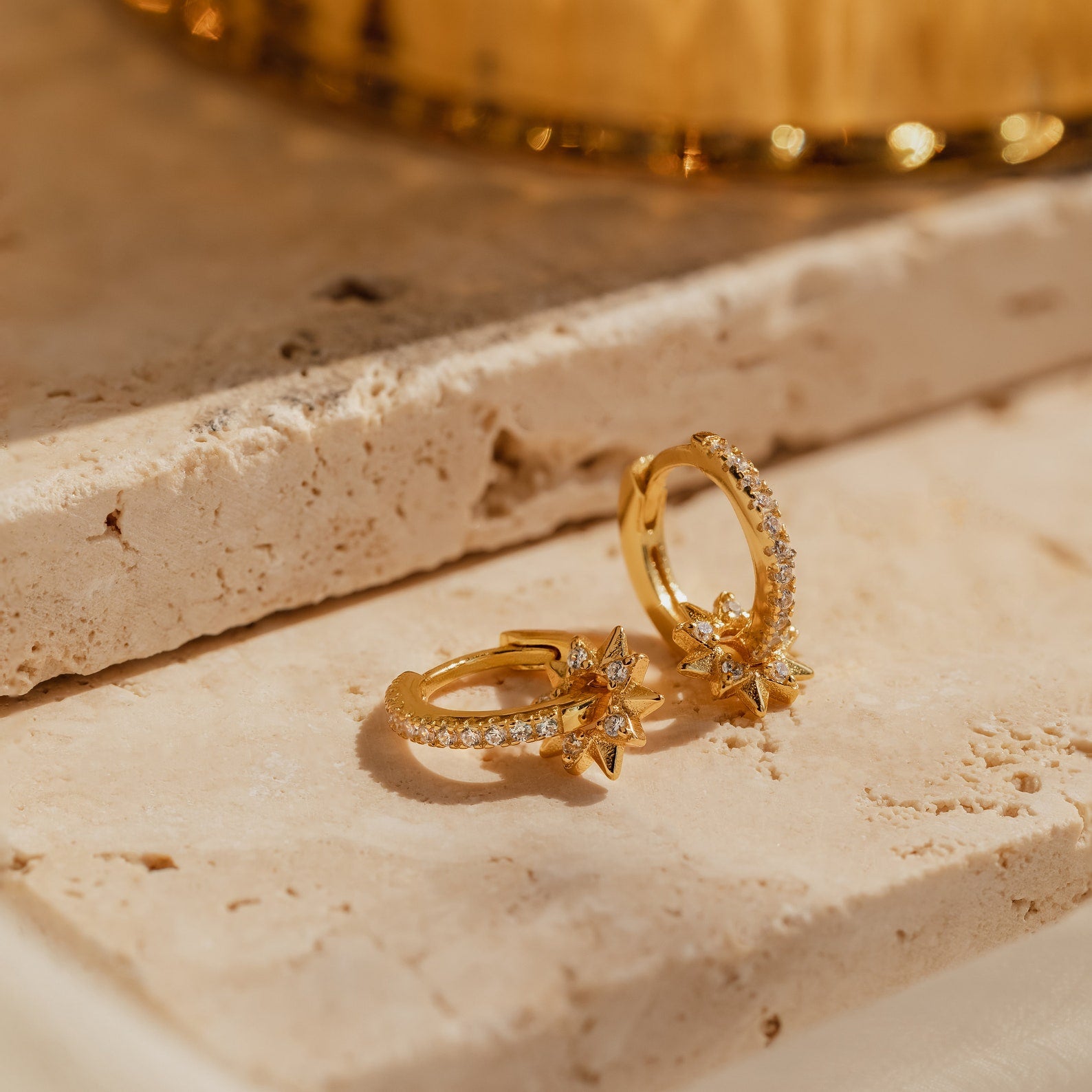 Two Pave Sun Huggies, gold hoops with small star-shaped charms and embedded crystals, rest on a beige stone surface.