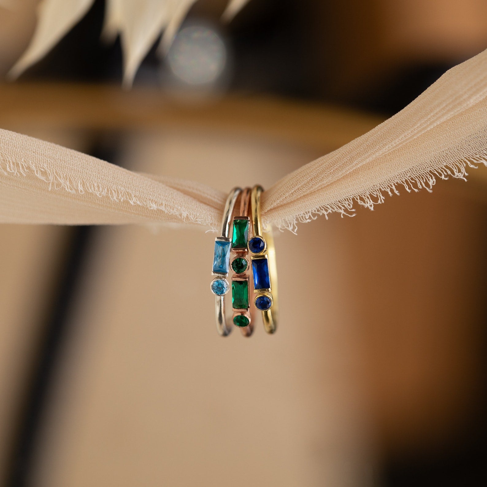 The Morse Code Birthstone Ring, featuring gold with blue and green gemstones, is showcased alongside two other rings on a soft beige fabric ribbon.