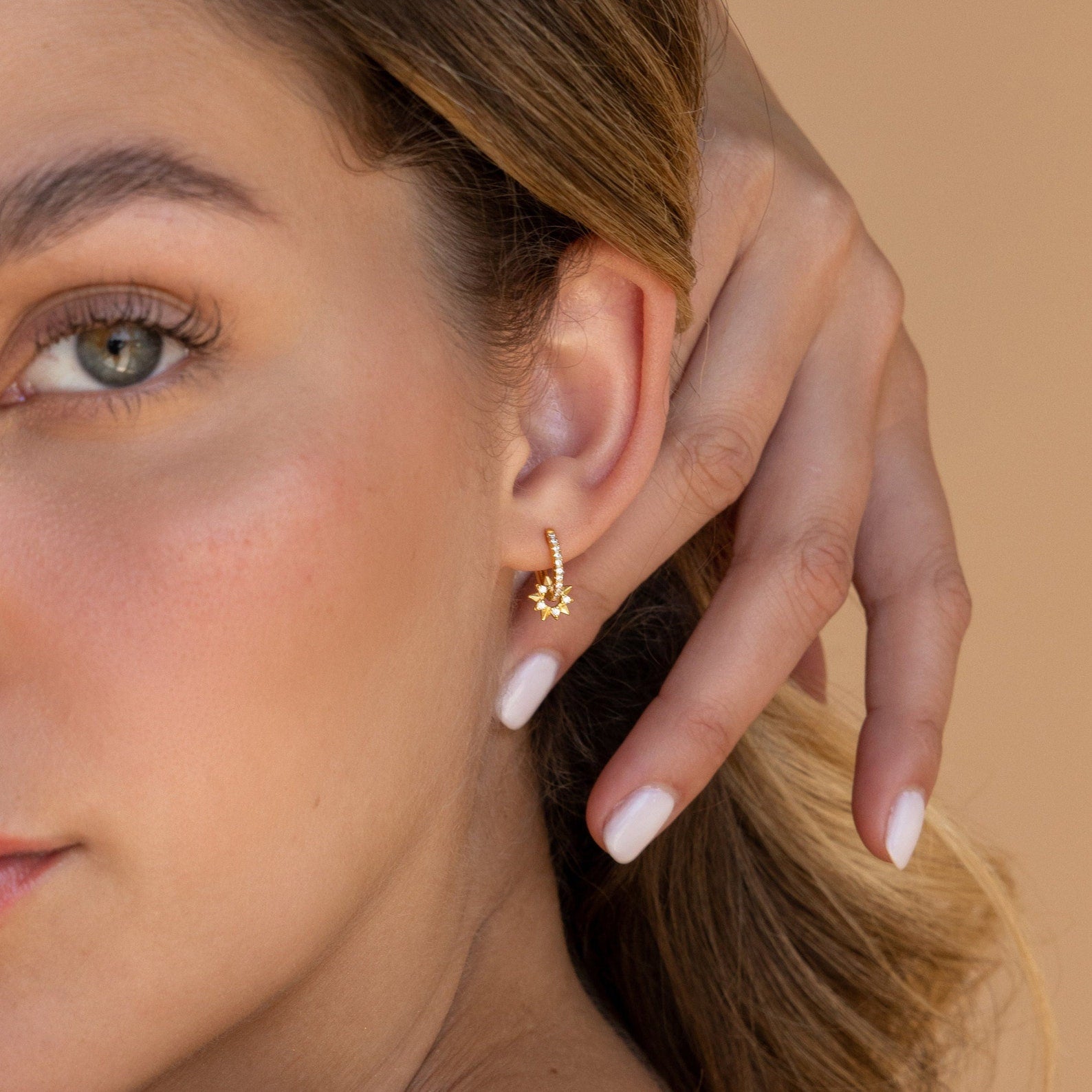 Woman with light skin wears Pave Sun Huggies, a small gold starburst earring, touching her ear against a neutral background.