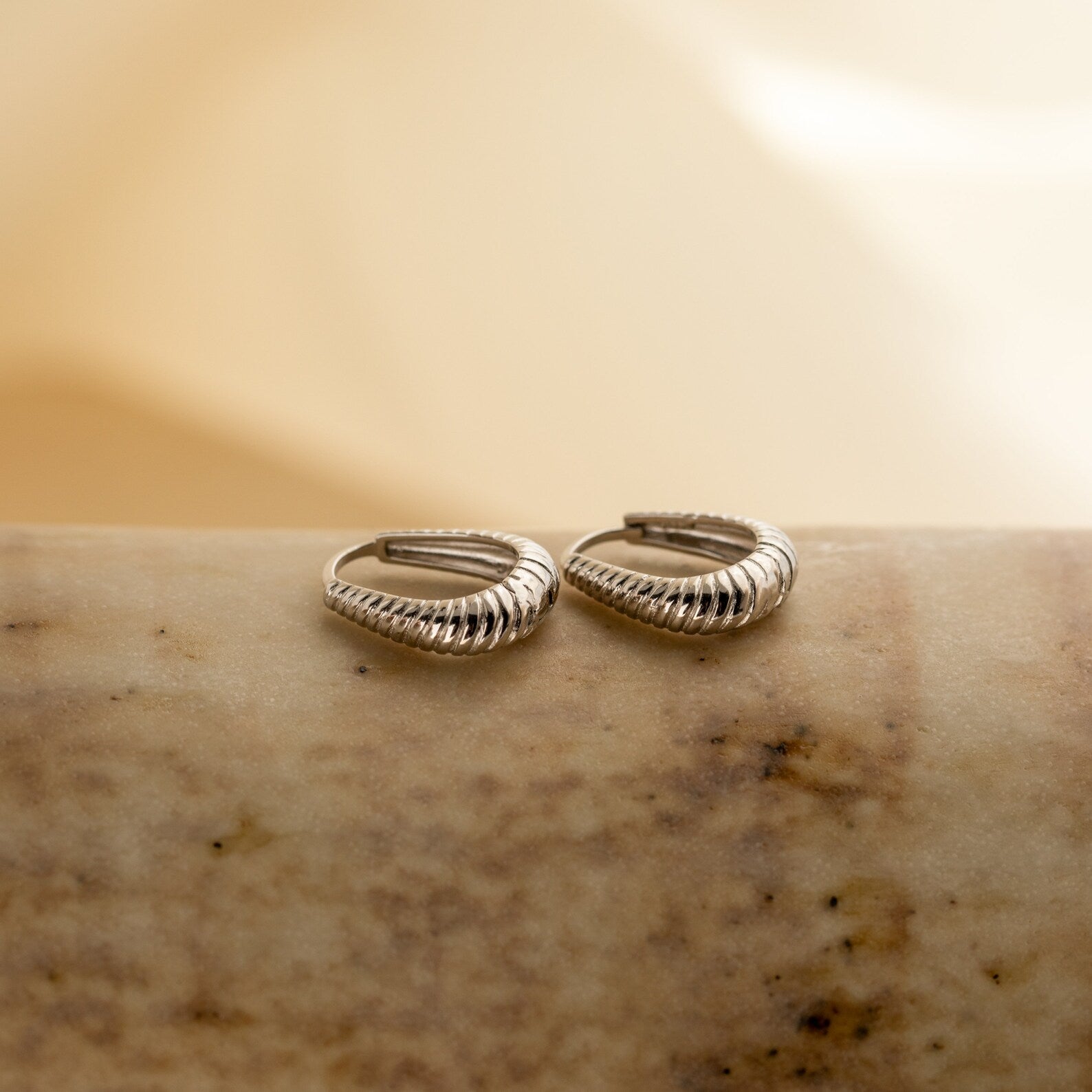 A pair of Bold Ribbed Hoops in silver with subtle geometric details are elegantly displayed on a smooth, marble-like surface.