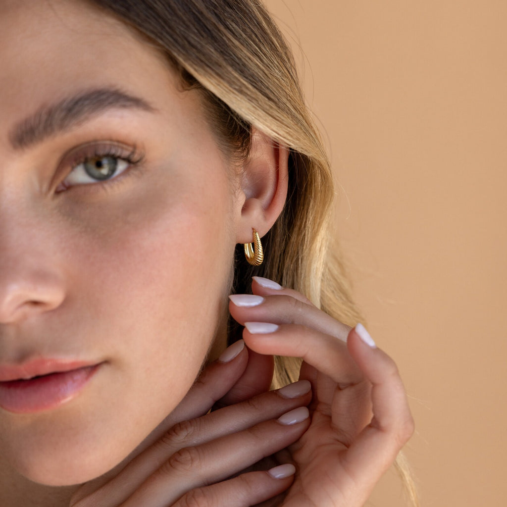 A woman with light eyes gently touches her ear, showcasing Bold Ribbed Hoops against a beige background.