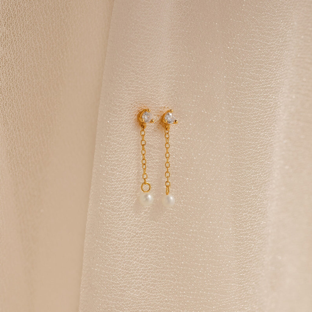 Diamond Pearl Drop Earrings with crescent moons, chain drops, and pearls are displayed on a soft beige fabric background, highlighting their timeless elegance.