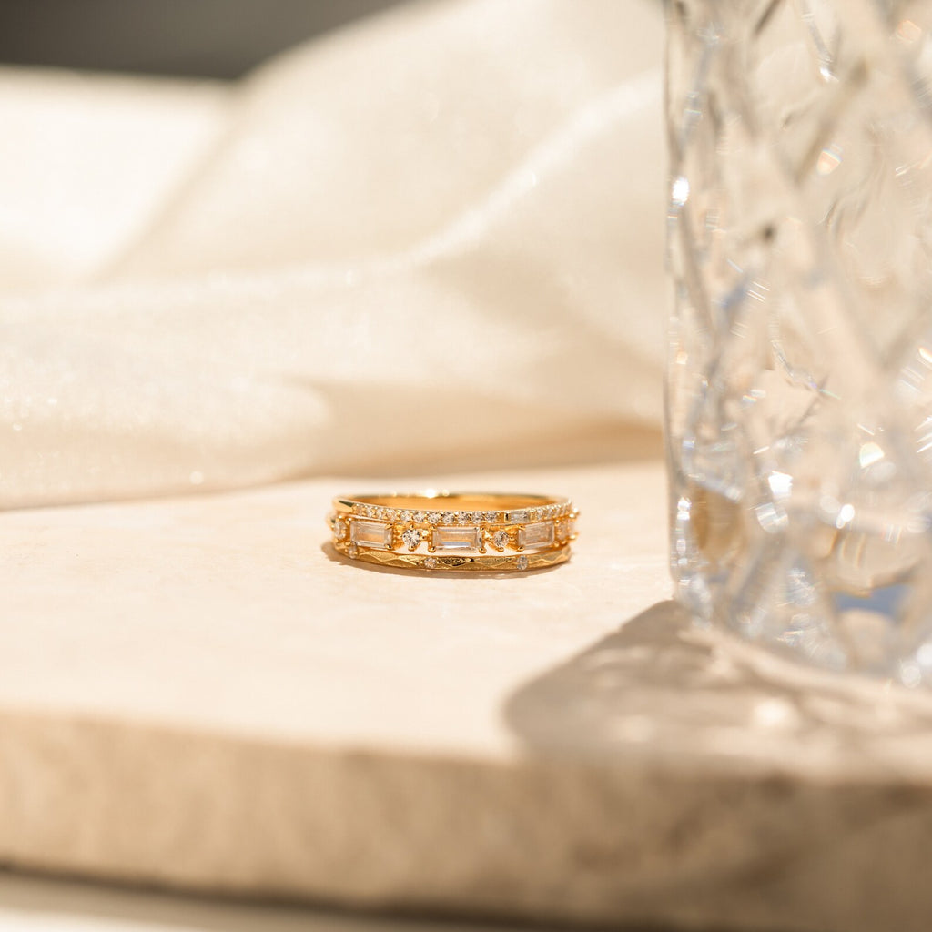 The Mote Diamond Stacking Ring Set in 18K Gold lies on marble beside clear textured glass and soft beige fabric, bathed in gentle sunlight—an ideal Christmas gift for someone special.