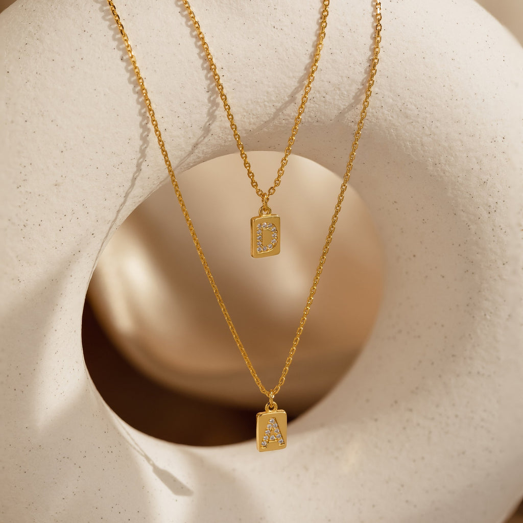Two gold Mini Pave Initial Tag Necklaces with D and A letter pendants are shown on a textured, cream sculpture—adding a unique touch of personalized jewelry.