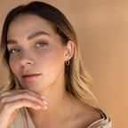 Woman with wavy blonde hair poses with hand near chin against a beige background, wearing the Medium Bold Hoops in 18K Gold for a stylish everyday jewelry look.
