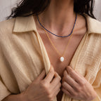 A woman in a beige shirt wears Boho Beaded Necklaces with gold, blue strands, and a large white pearl pendant—an ideal accessory to elevate summer outfits.