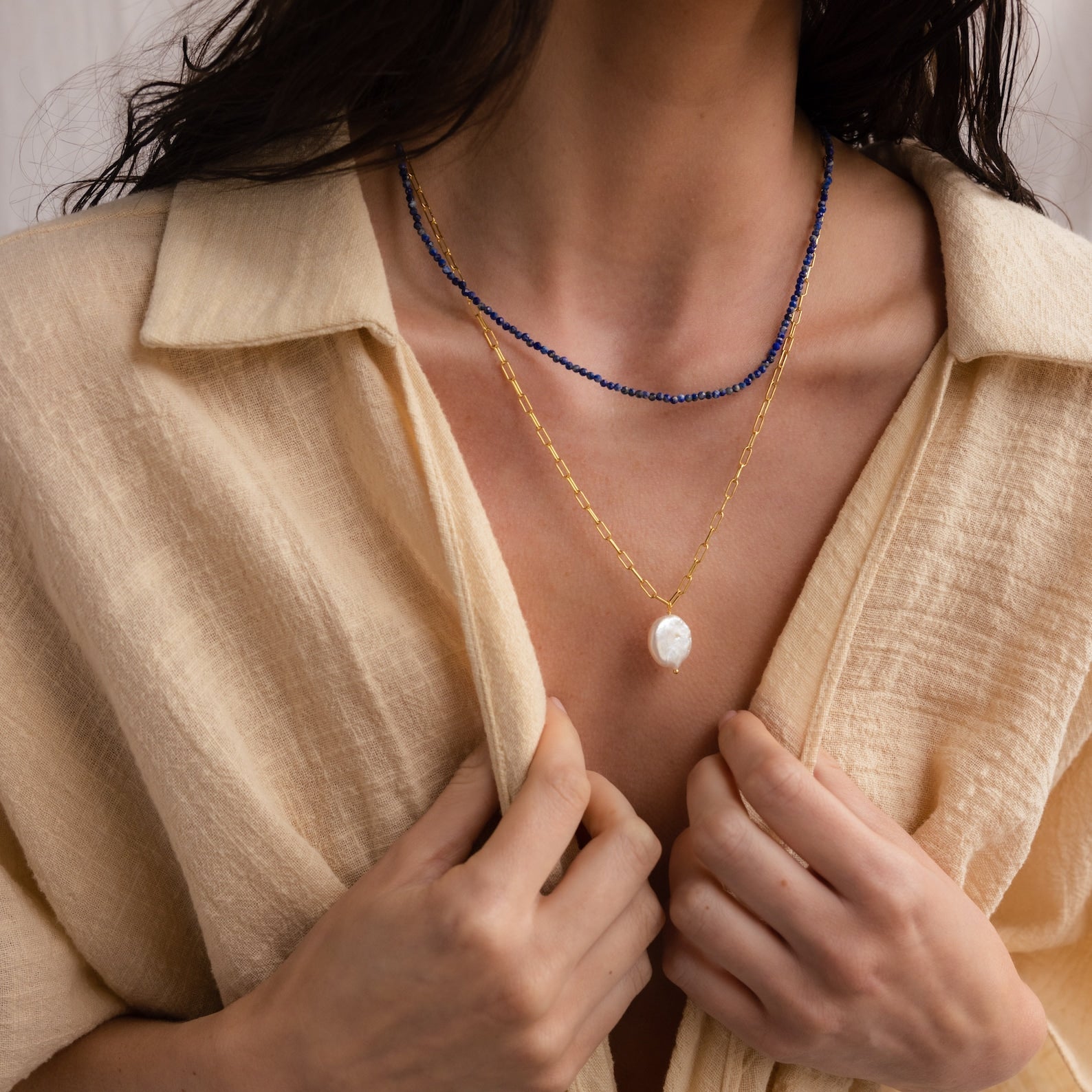 A woman in a beige shirt wears Boho Beaded Necklaces with gold, blue strands, and a large white pearl pendant—an ideal accessory to elevate summer outfits.