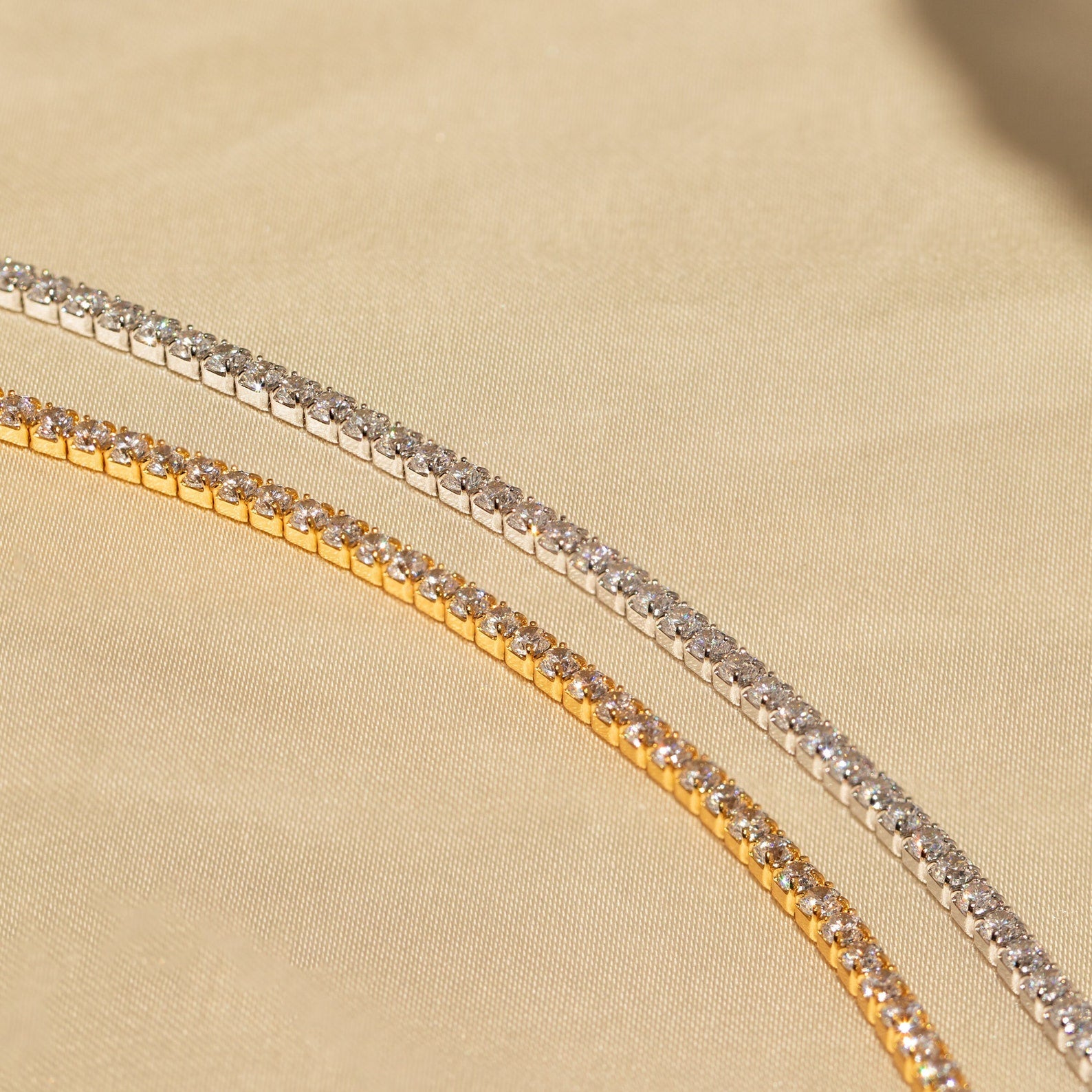 The Classic Diamond Tennis Bracelet, available in gold and silver, features sparkling gemstones and a timeless design—an elegant jewelry piece displayed on a beige fabric background.