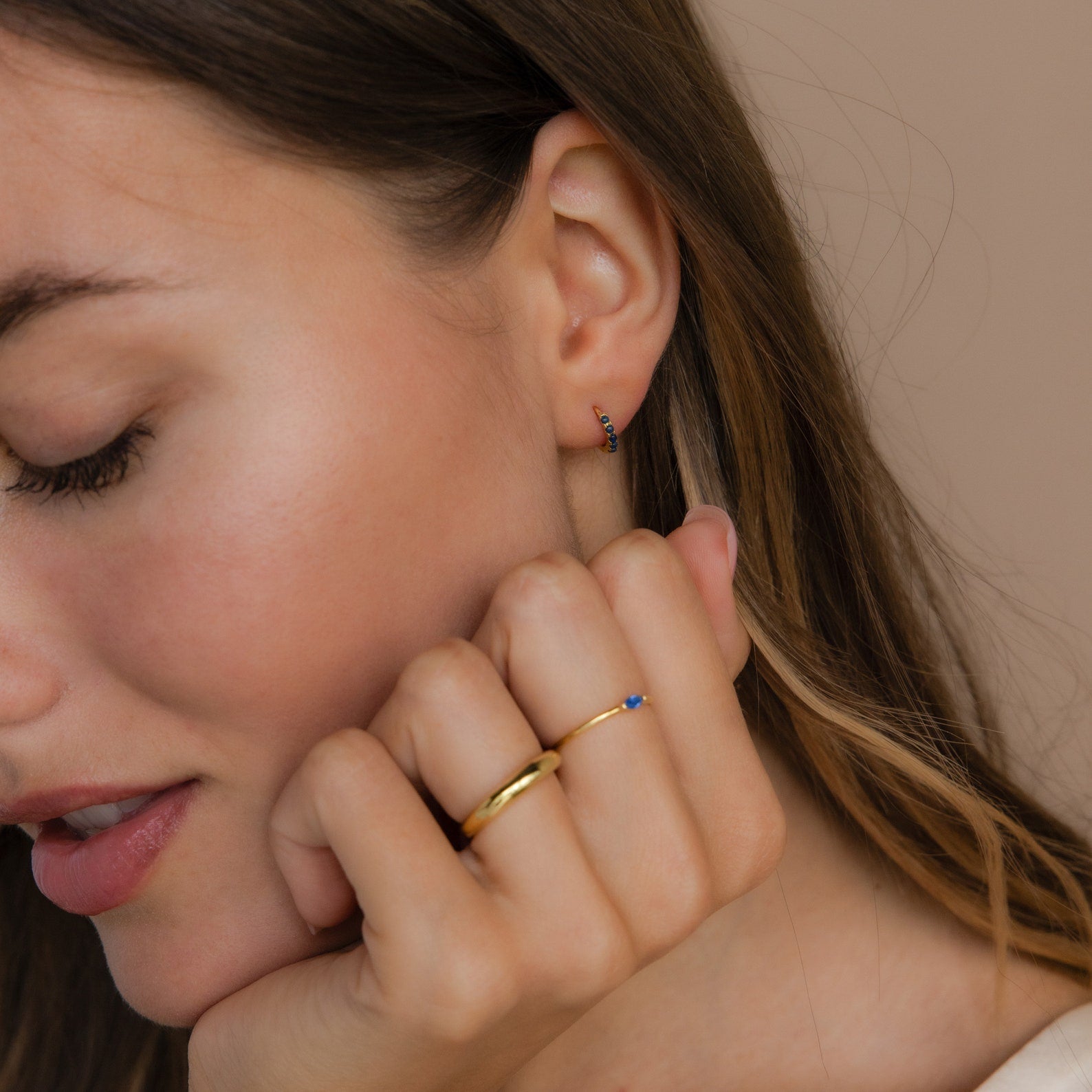 A woman with closed eyes wears a gold ring and Indigo Blue Opal Huggies, adding a minimalist touch to her look.