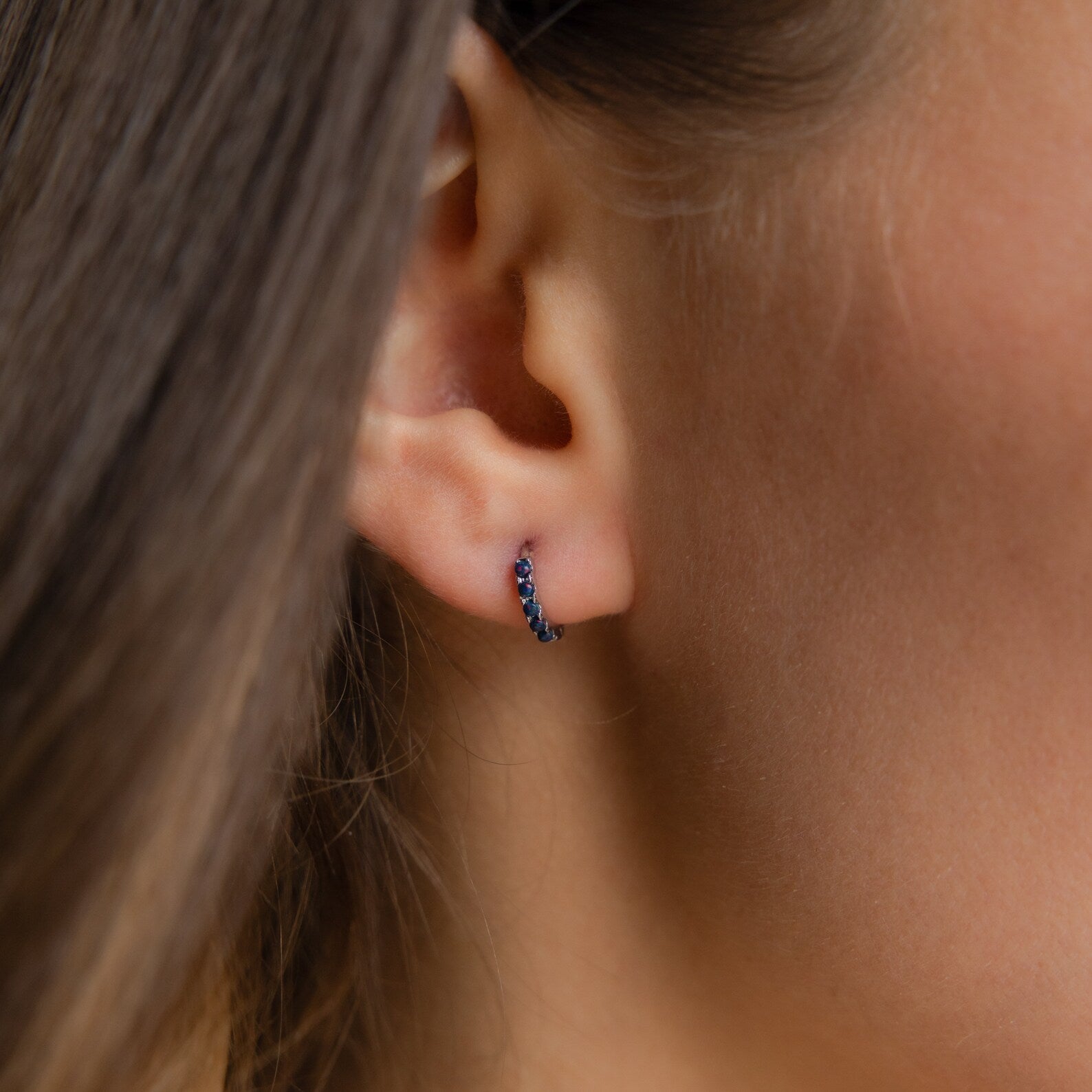 Close-up of a woman's ear wearing Indigo Blue Opal Huggies, minimalist hoop earrings adorned with indigo blue opal stones.