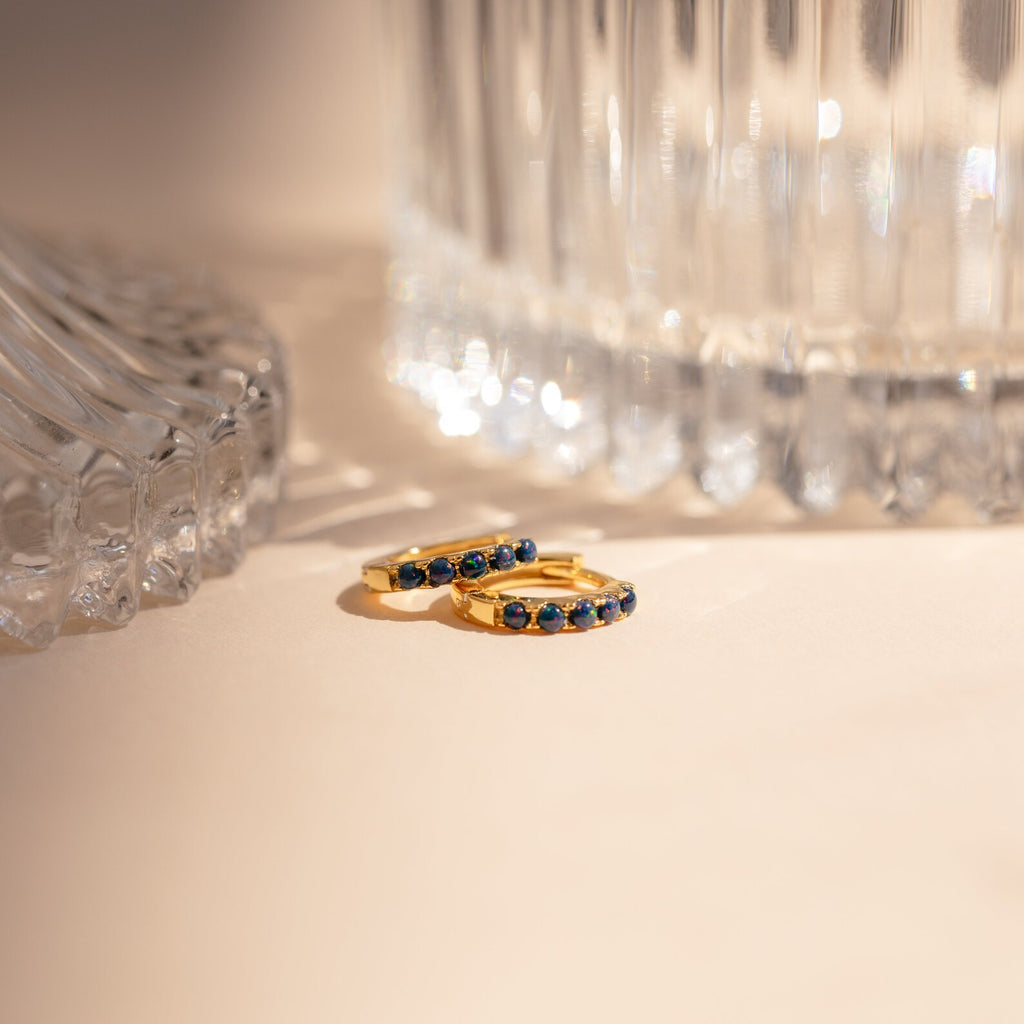 Indigo Blue Opal Huggies—gold hoop earrings with blue opals—rest on a beige surface beside clear glass vases in soft light, offering a minimalist and versatile style perfect for any occasion.