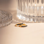 Indigo Blue Opal Huggies—gold hoop earrings with blue opals—rest on a beige surface beside clear glass vases in soft light, offering a minimalist and versatile style perfect for any occasion.