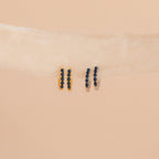 Two pairs of small, minimalist earrings—gold and silver Indigo Blue Opal Huggies with dark stones—are displayed on a light beige ribbon.