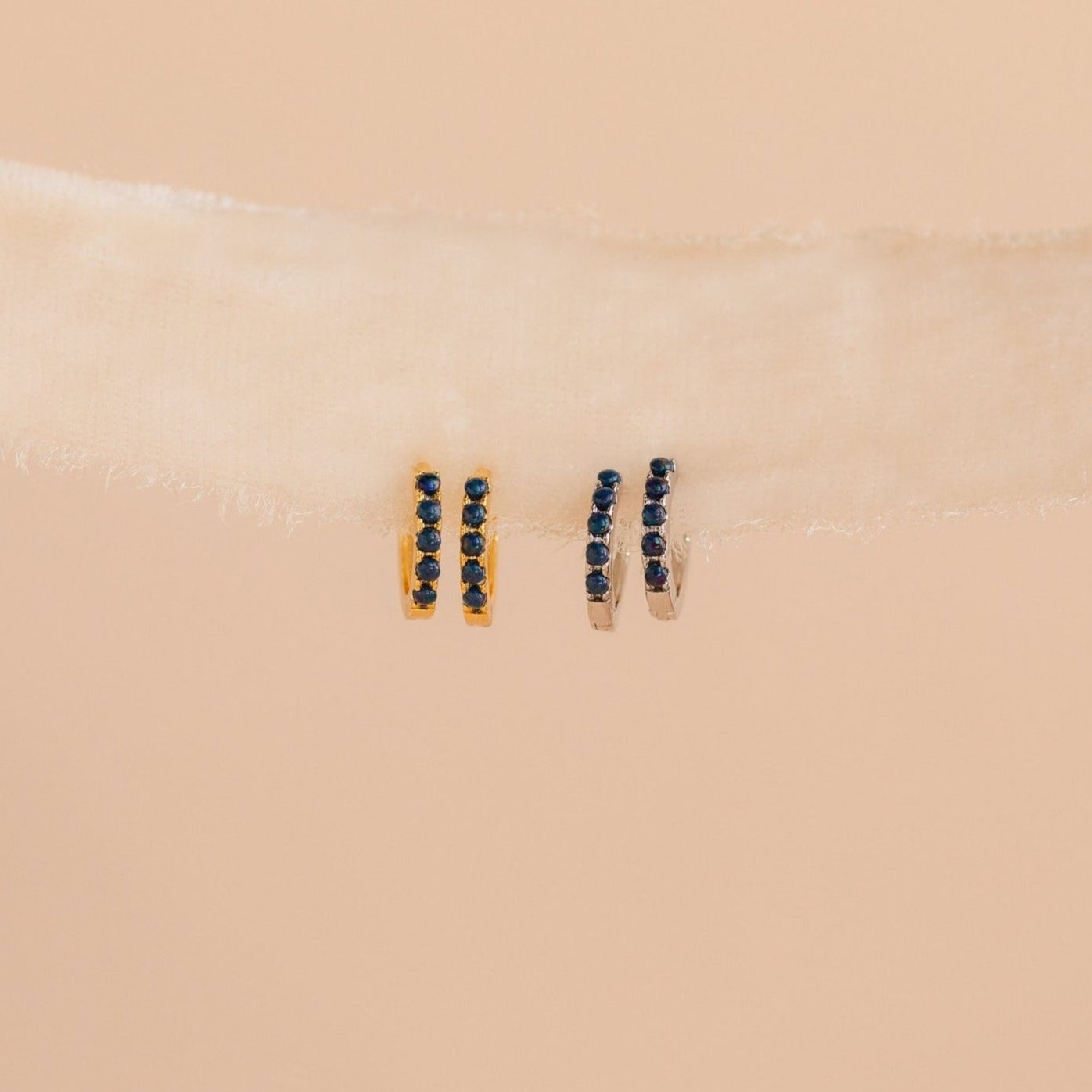 Two pairs of small, minimalist earrings—gold and silver Indigo Blue Opal Huggies with dark stones—are displayed on a light beige ribbon.