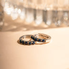 Two Indigo Blue Opal Huggies, featuring silver bands with indigo blue opal hoops, rest side by side on a light surface, showcasing their minimalist elegance against a softly blurred background.