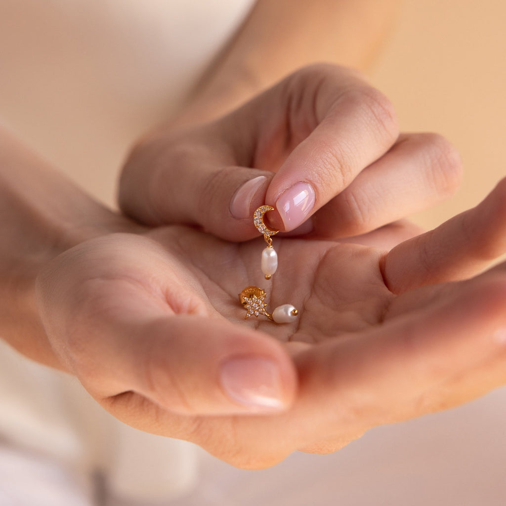 A hand holds a delicate gold earring with a pearl charm and small star, from the Moon & Star Pearl Earrings collection.