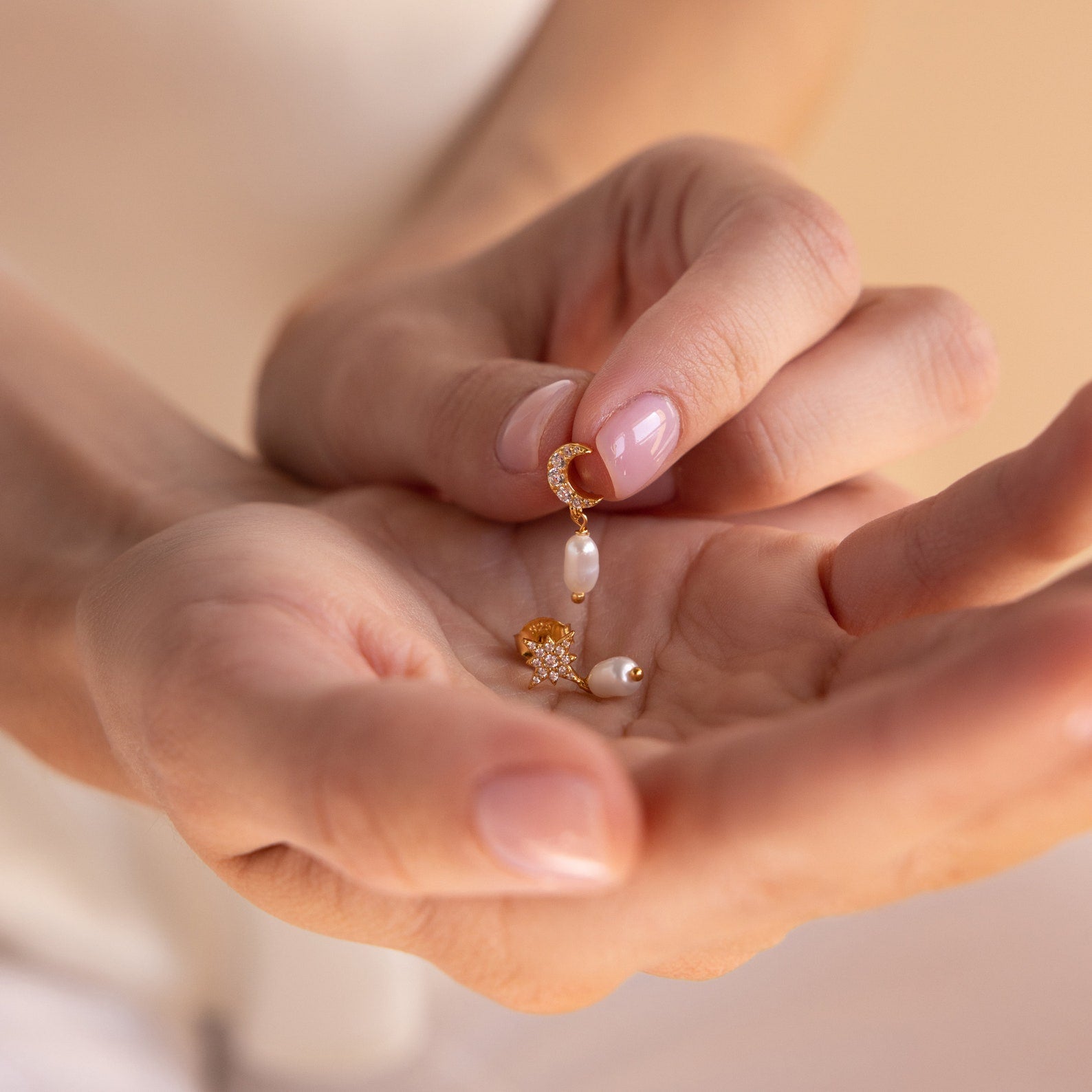 A hand holds a delicate gold earring with a pearl charm and small star, from the Moon & Star Pearl Earrings collection.