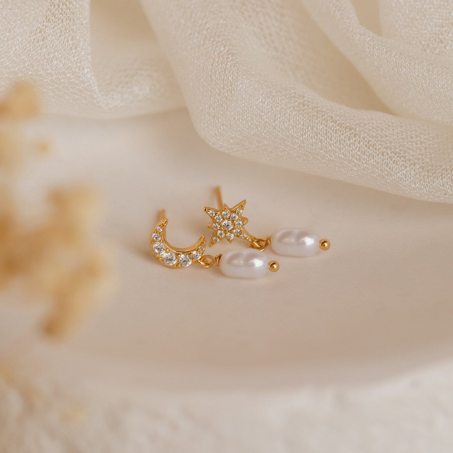 Moon & Star Pearl Earrings, featuring crystals, pearls, and a delicate pearl charm, shine beautifully against a soft beige fabric background.