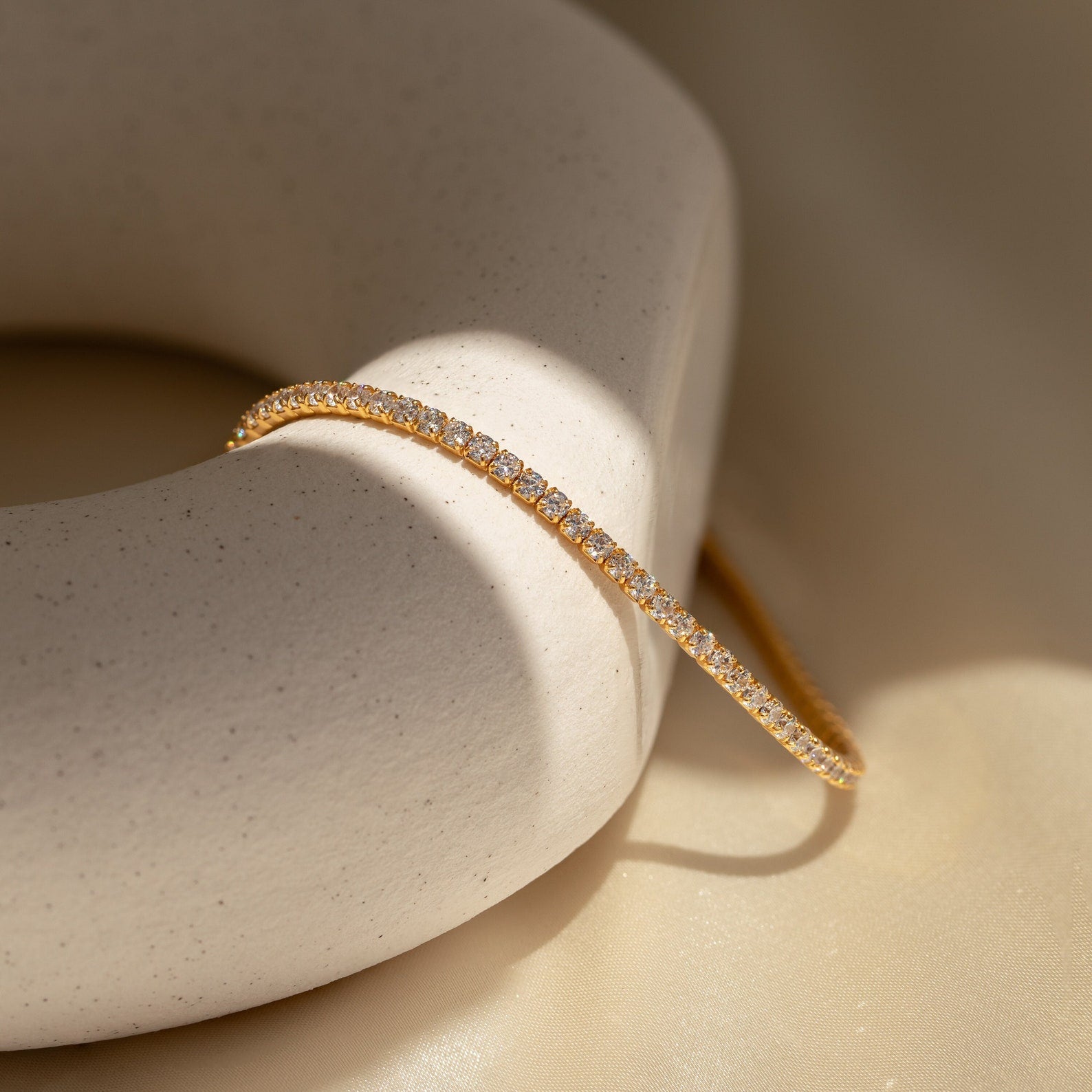 The Classic Diamond Tennis Bracelet in 18K Gold rests on a round, off-white stone, beautifully illuminated by soft natural lighting.