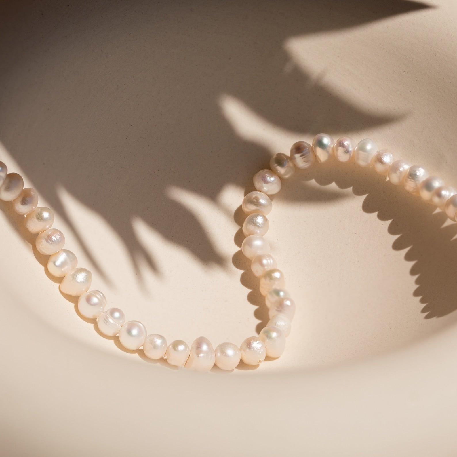 A string of irregular white pearls forms elegant Beaded Belly Chains on a light beige surface, with soft leaf shadows adding a touch of bohemian charm.