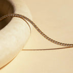 The Tennis Necklace Set features a gold tennis bracelet with diamonds on a cream-colored curved surface, ideal for layering with the diamond tennis necklace to create a sophisticated, elegant look.