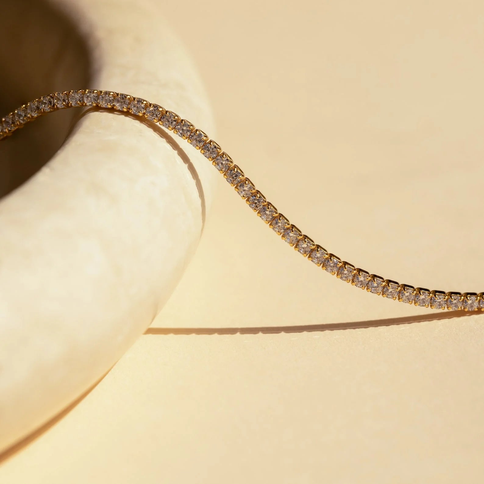 The Tennis Necklace Set features a gold tennis bracelet with diamonds on a cream-colored curved surface, ideal for layering with the diamond tennis necklace to create a sophisticated, elegant look.