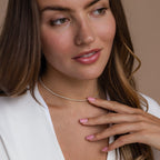 A woman in a white top softly touching her gold diamond tennis choker necklace, with a serene expression and pink manicured nails.
